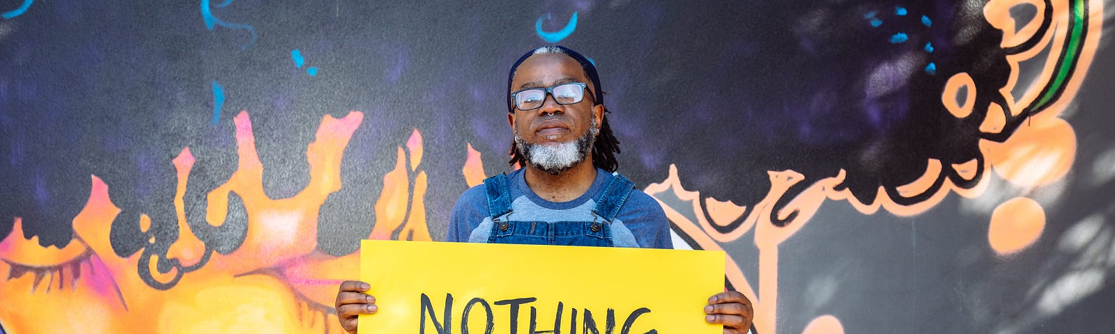 A Deaf Black man with a septum piercing and a black and white beard, wearing denim overalls and glasses, holds a hand-lettered sign that reads ‘NOTHING about us without US.’ A vibrant mural of a Black woman serves as the background.
