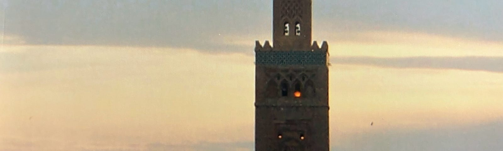 A Moroccan tower towering above the trees with sky the colors of yellow and orange as the sun set.