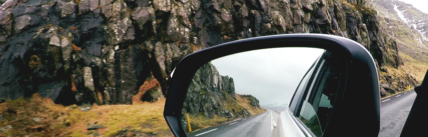 The rearview mirror of a car showing the open road. In the background are tall, rocky mountains and green hills.