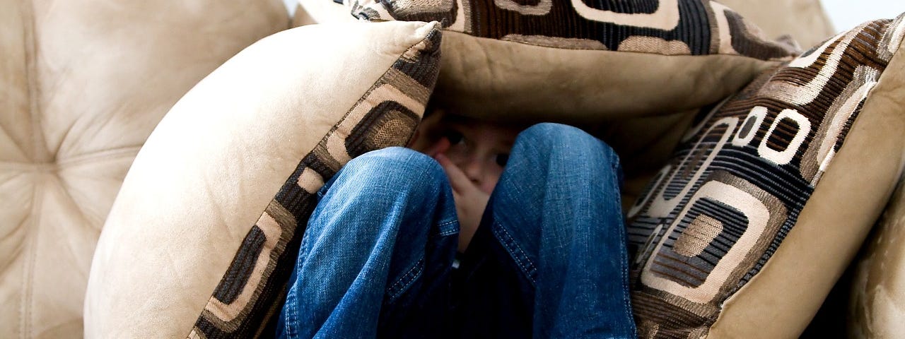 A child wearing blue jeans lying on a beige couch and buried among beige-brown geometric-patterned cushions.