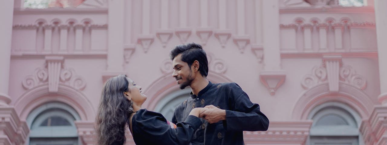 A young South Asian couple dance in front of a beautiful pink building.