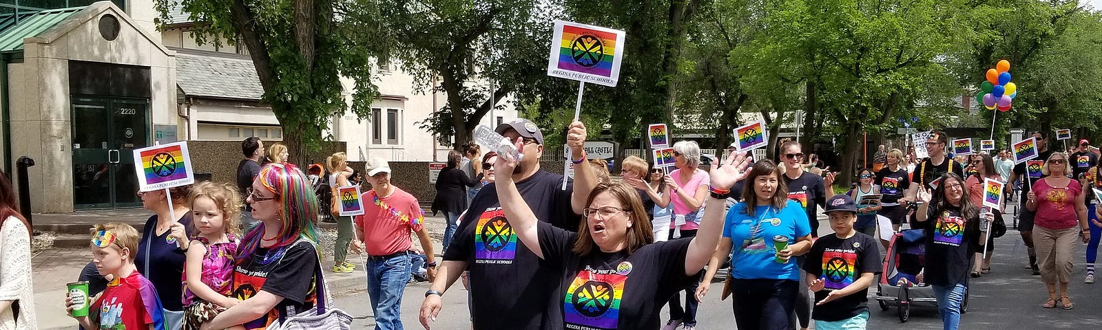 A group of school staff and children march in a Pride parade. Most are wearing black shirts with rainbow logos and some are carrying rainbow signs. One adult has rainbow-dyed hair and is carrying a small child wearing a rainbow tutu.