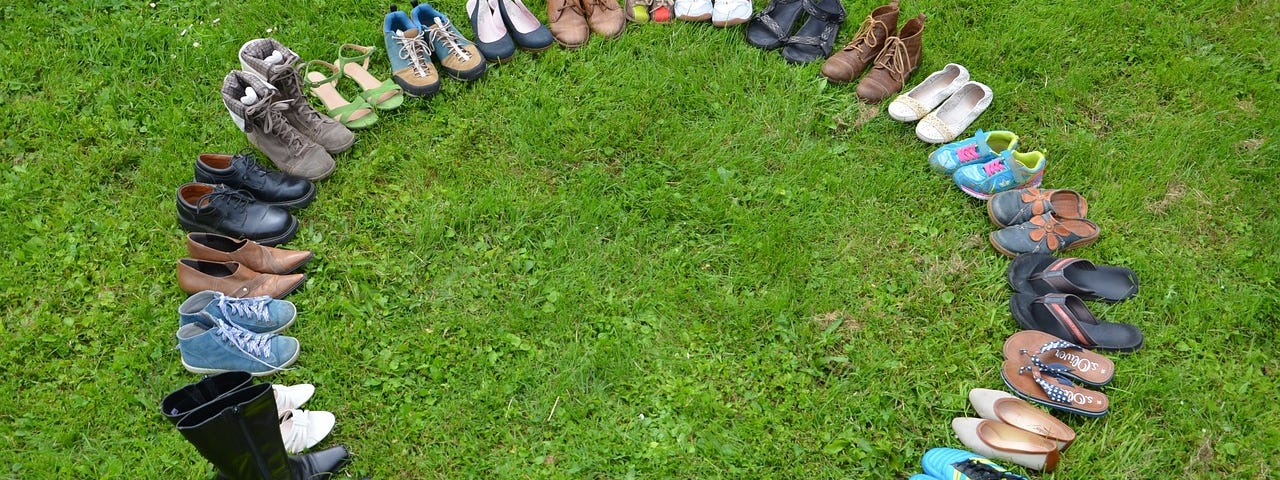 27 pairs of varied styles and sizes of footwear in a circle on grass.