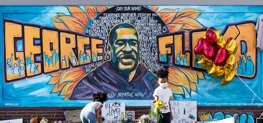 An image of a mural of George Floyd with people setting flowers and signs beneath it.