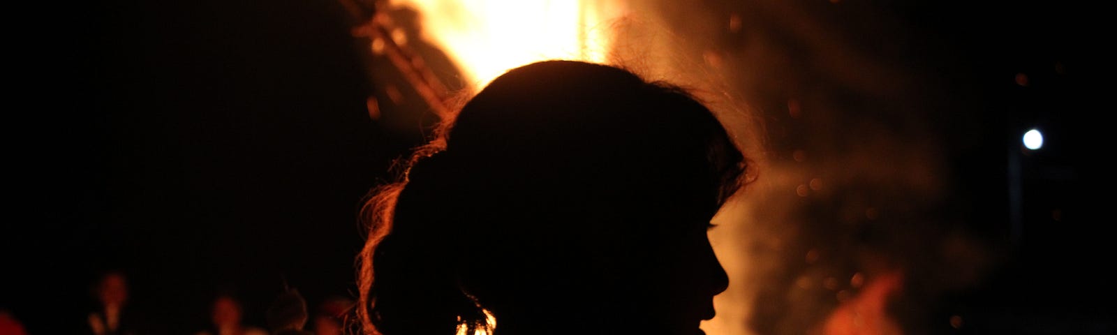 The silhouette of a woman sits against a roaring fire at night.