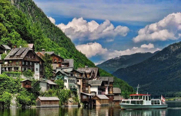 Old wooden German homes of brown cling to green slopes by the Rhine River with mountains and a boat in the background and foreground.