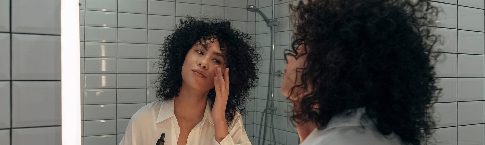 Woman in a light cotton shirt and curly dark hair applies skin serum in a subway-tiled bathroom mirror