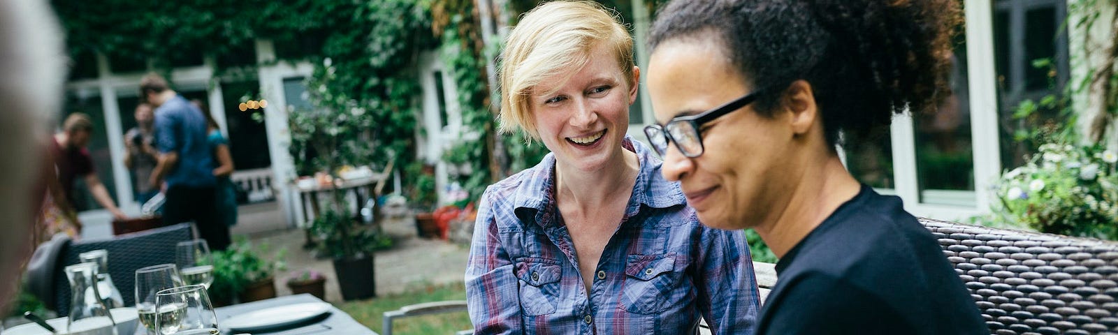 At a table set for a meal, a white woman with a kindly smile looks over at a woman of color with a slight grimace