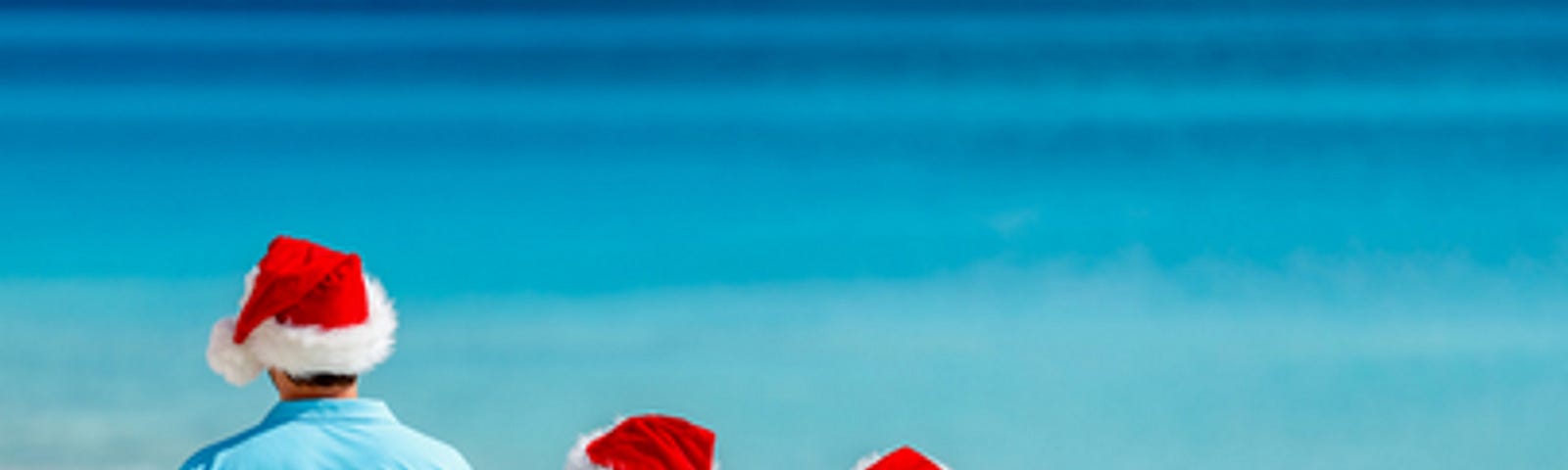 Father and two kids on beach wearing santa hats and watching the blue ocean.