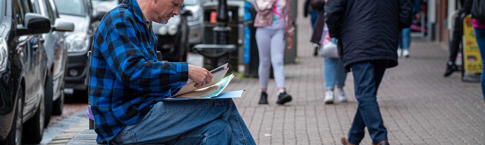 Rick, a middle aged white man, wearing a blue and black checked shirt and blue jeans, sitting on a black bench looking through an activity pack. People and signs on the High Street behind him.