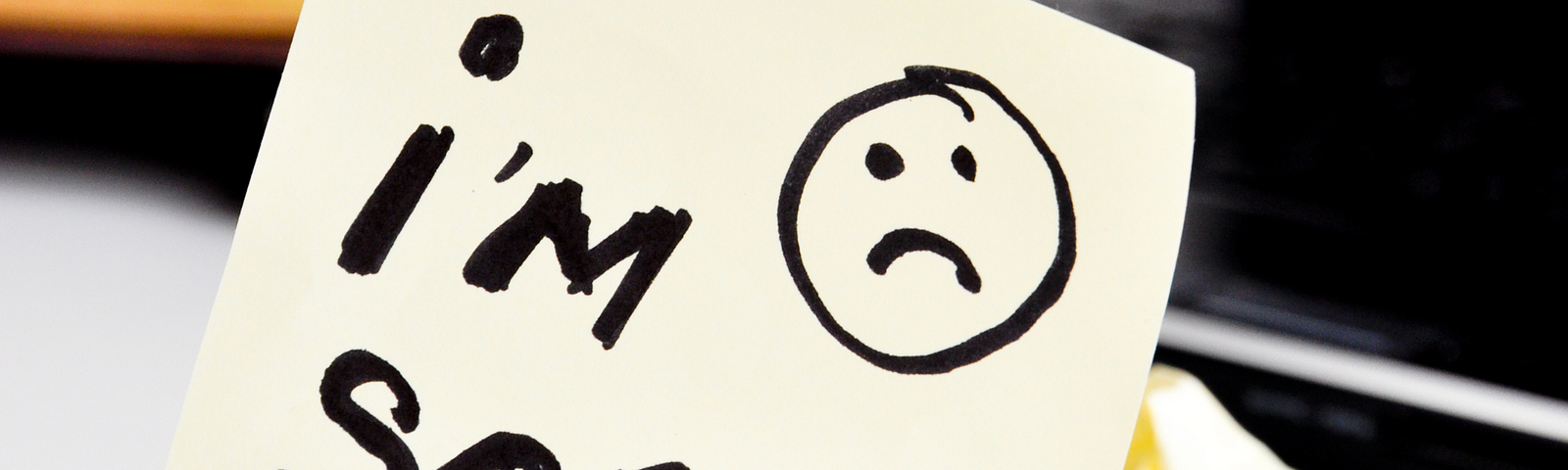 This image shows a yellow sticky note with the handwritten message “I’m Sorry…” accompanied by a sad face drawing. The note is placed on a desk near a keyboard and some crumpled paper.