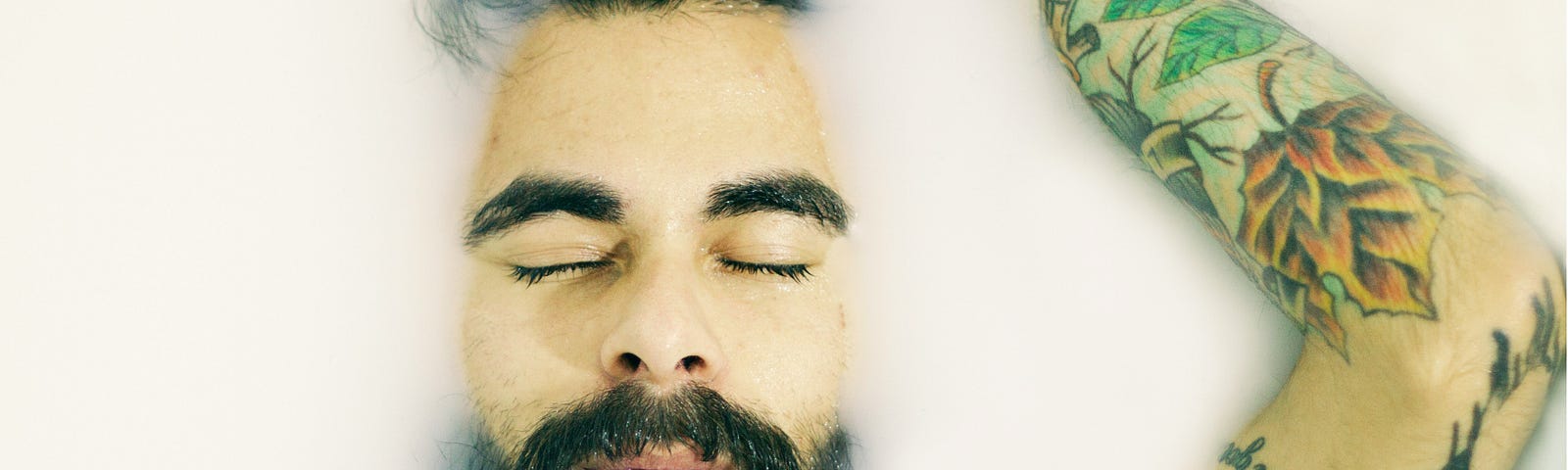 A tattooed and bearded man rests his eyes in a milk bath.
