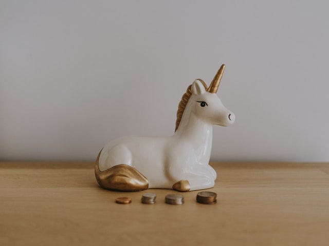 Ceramic white unicorn with a gold horn sitting in front of stacks of coins.