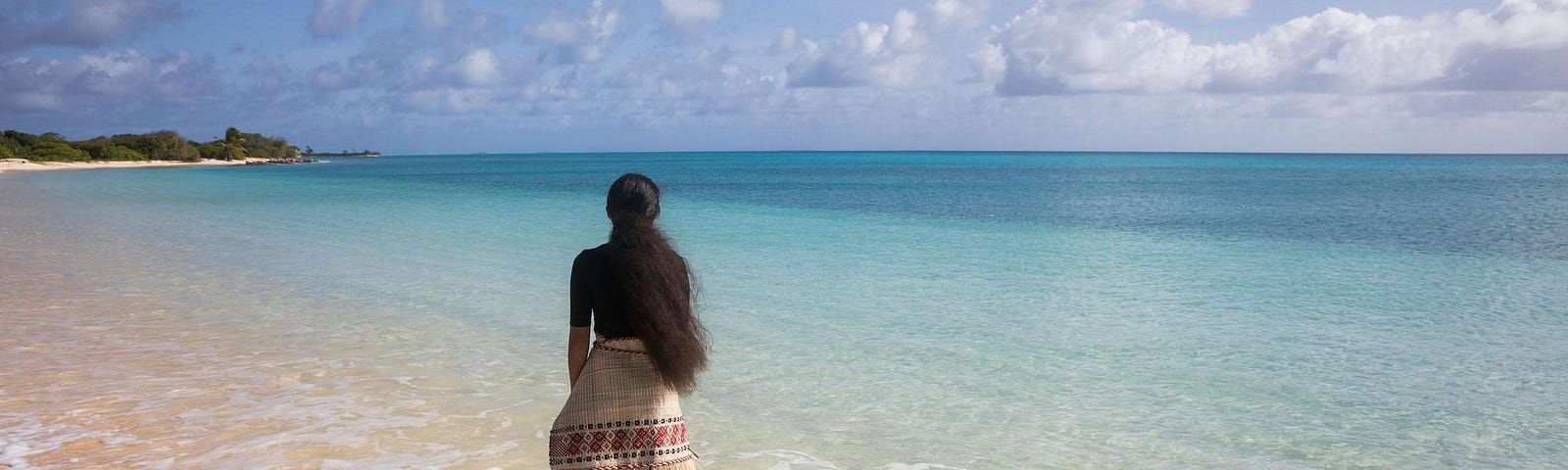A woman, Kathy Jetñil-Kijiner, stands at the shoreline with her back turned to the camera. She is wearing a traditional jaki-ed. The sky is filled with clouds and the ocean is clear and crystalline.