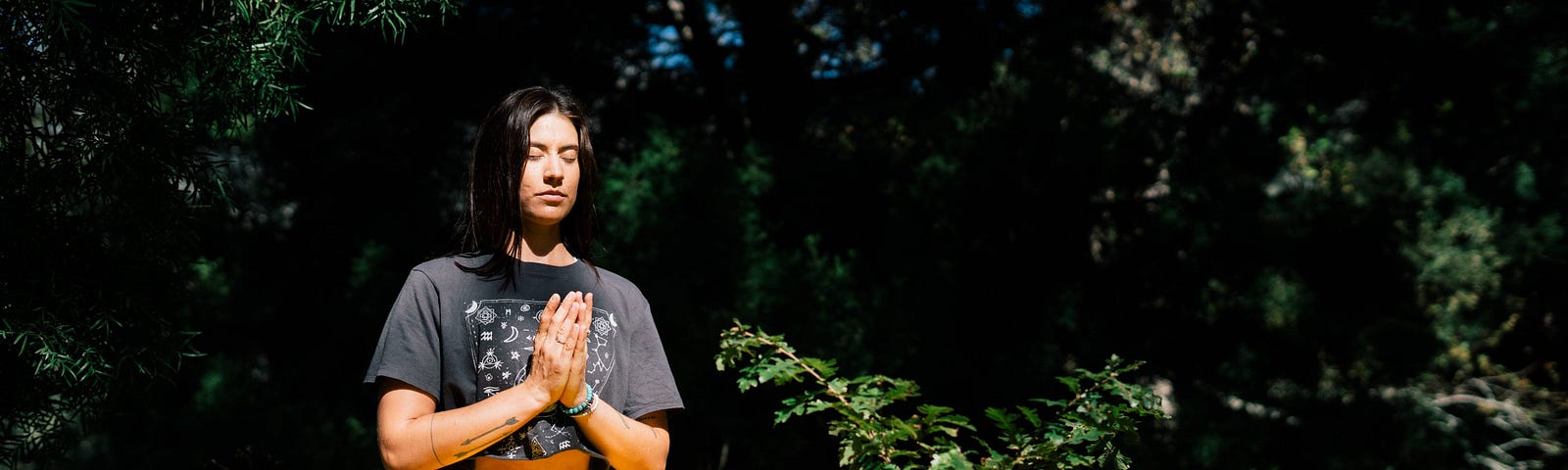 A young, dark haired woman in grey crop top and yellow leggings sitting cross legged on a rock, meditating with her palms together. She is surrounded by greenery.