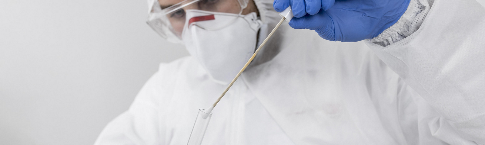 A person wearing a mask, gloves, and protective clothing inserting a nasal swab into test tube.