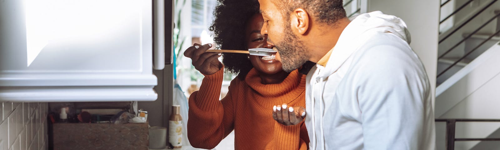 A man and woman are cooking in their kitchen, and the woman is giving him a bit of food to taste.