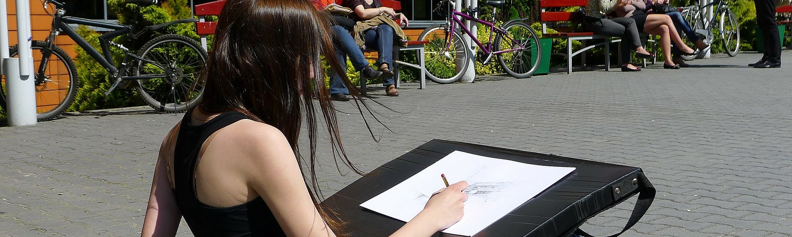 A young woman sketches outdoors, sitting on the ground. She is wearing a black tank top, jeans, and red sneakers