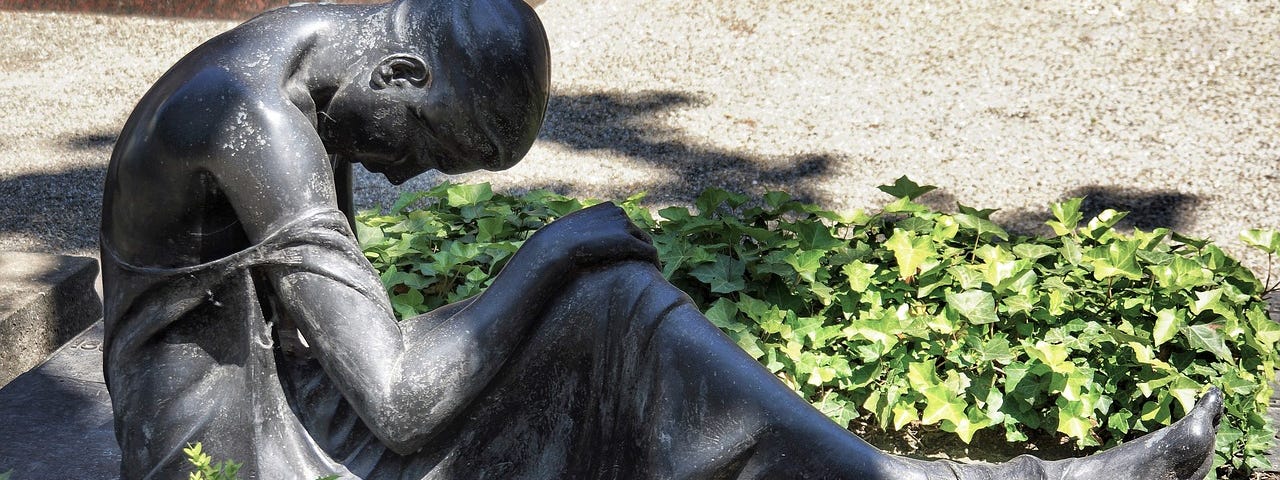 A statue of a woman with her head hung. She is laying in greenery, but it appears she is in a cemetery.