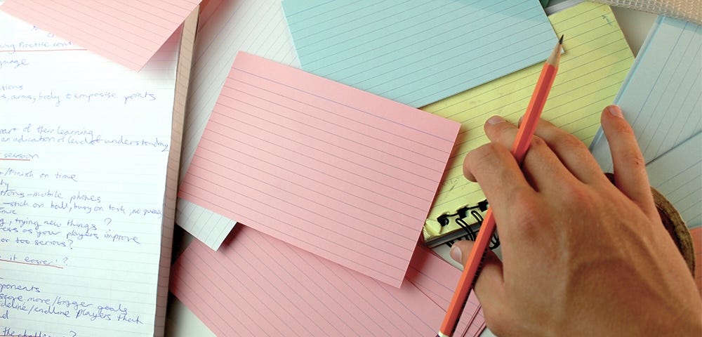 A notebook and pink and blue notecards on a table. Someone’s hand holds a pencil and reaches for a yellow notebook.