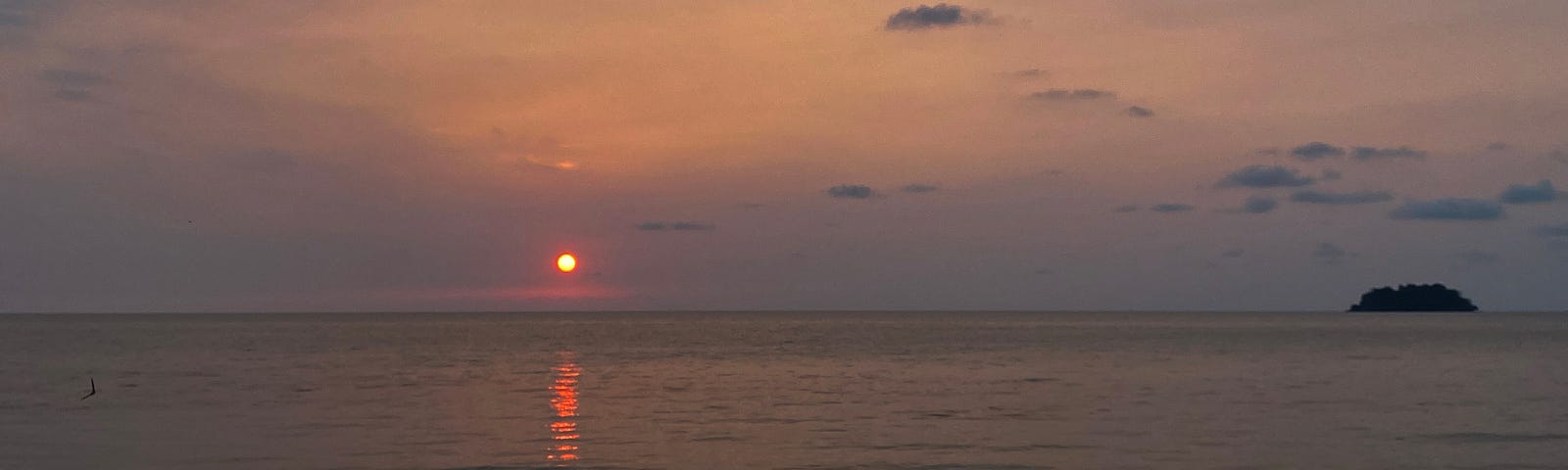 Sunset over the sea. Small orange sun is almost at the horizon, reflected in the water. A small island is on the right side of the photo on the horizon. A rocky shore is in the foreground.