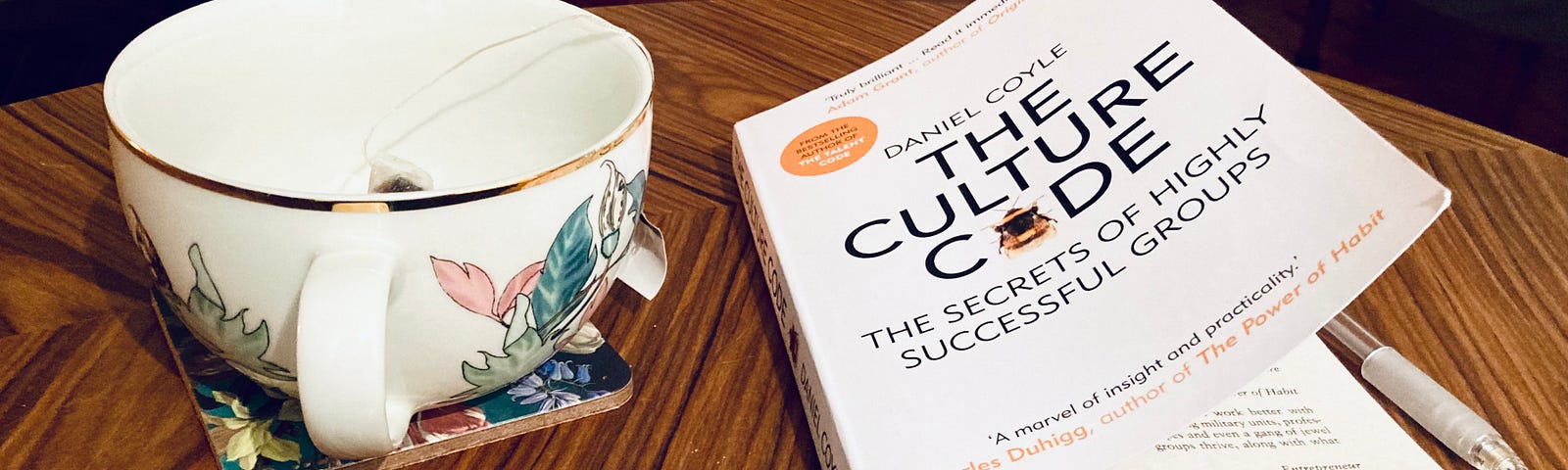 A dark wood coffee table. On the left, a white flowery tea mug, to the right a copy of the book “The Culture Code”