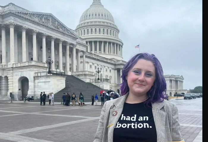 A photo of Lizzie, a White woman with purple hair. She is wearing a T-shirt that reads “had an abortion,” and holding a sign that says “ABORTION JUSTICE NOW. The Capitol building is in the background.