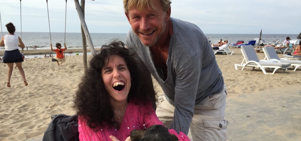 A blond man leaning over a long-haired woman in a wheelchair holding a little dog. They are on the beach.