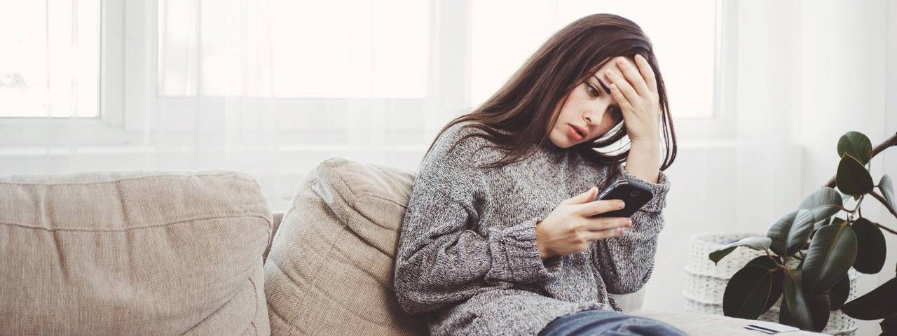 A woman sits on a couch looking at her cellphone with her hand on her forehead, looking frustrated and distressed.