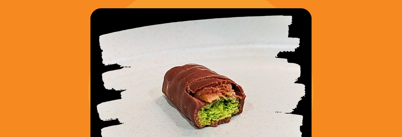 Half of Twix candy bar showing green cookie interior. Photo appears in light-colored “paintbrush” frame, which is in turn on a black background and then a larger orange background.