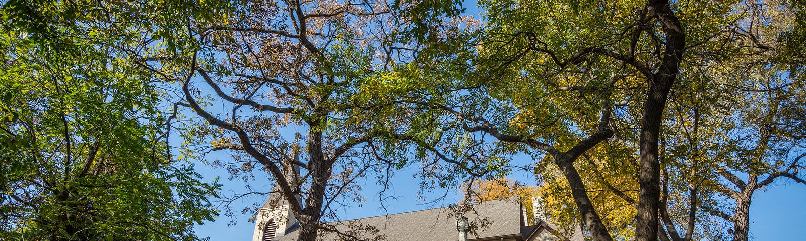 A view where one is looking up from the base of a hill at a classic church with steeple. Trees and graves line the hill.