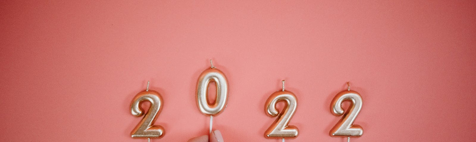 A white hand puts gold candles spelling out “2022” on a pink background.