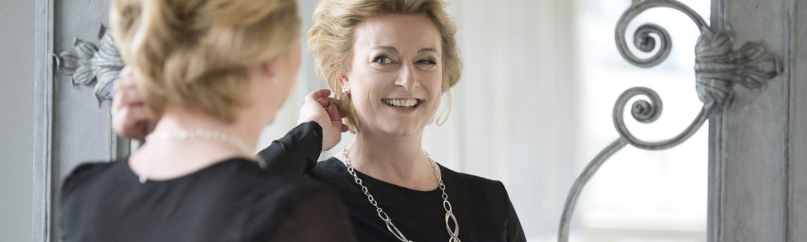 A mature woman with blonde hair wearing a black dress with long sleeves and a long silver chain smiling at herself in a mirror.