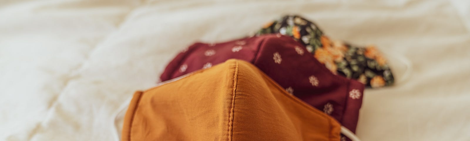 Three fabric face masks — one yellow, one maroon with yellow flowers, and one black with big yellow roses — stacked on a bedcover.