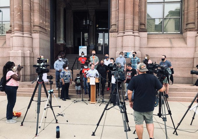 Jack Jose stands at the podium on the steps of Cincinnati City Hall, surrounded by Neighborhoods United supporters and media.
