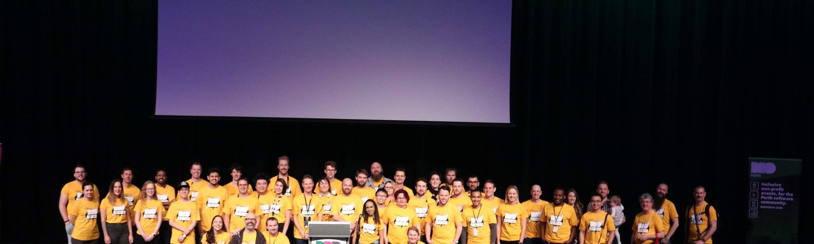 Over 50 people on a large stage all wearing yellow shirts that say “DDD Perth 2019 Team”. Some people in the front are kneeling. The majority of people are standing in multiple rows. There is a lectern in the front with a DDD Perth banner on it.
