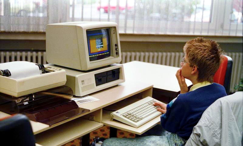 A boy, of about 8 years old, is sitting at a desk in front of an old IBM computer. There is a dot matrix printer on his left. The computer includes a 5.25 in floppy disk drive as well as a 3.5 in floppy disk drive.