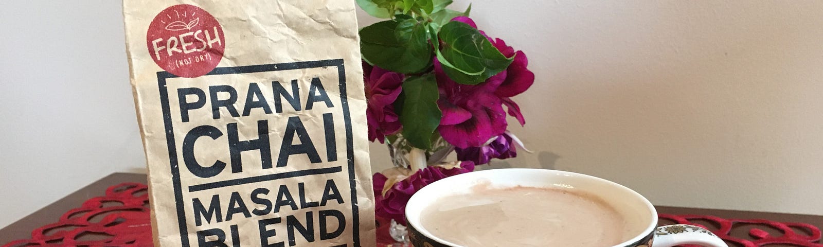 A paper packet labeled ‘Prana Chai Masala Blend’ next to a cup of beige liquid set on a table with a red cloth and flowers.