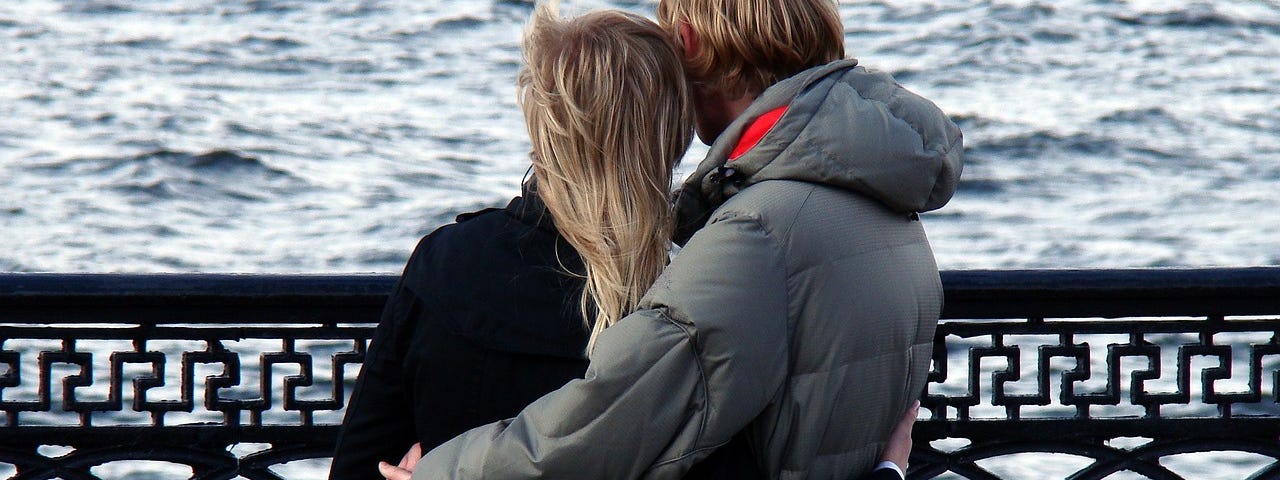 Two people with backs to camera, arms around each other, wearing coats, looking out over rough water.