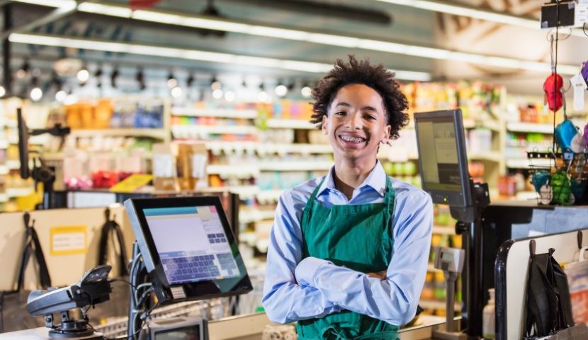 A student poses for a photo behind a supermarket cash register.