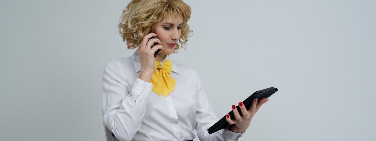 Image shows a blond woman wearing a short black skirt, black stockings and a white blouse with a yellow bow, holding a phone to her ear with her right hand, and a tablet in her left hand. She’s looking at the tablet.