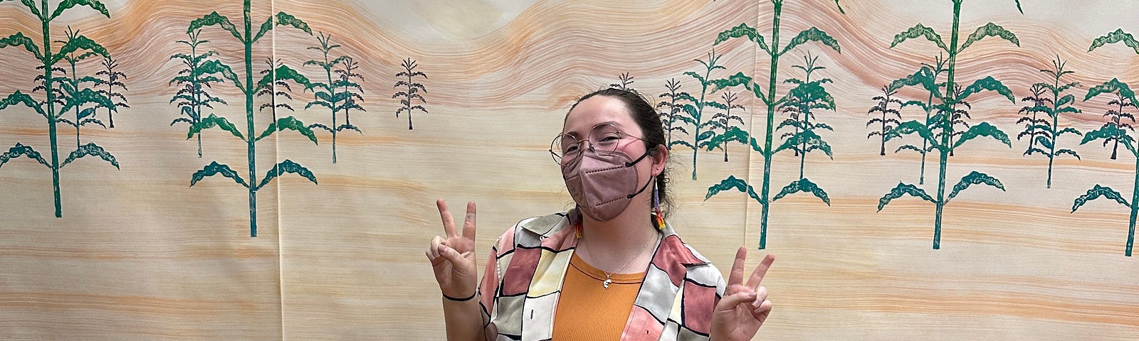 Theo Gardner wearing a terracotta checkered shirt, Yellow top, black pants, converse, and a brown mask. Posing in front of their art piece about Dinétah, the Navajo (or Diné) ancestral homeland. The art is a landscape with moutnans and Corn stocks