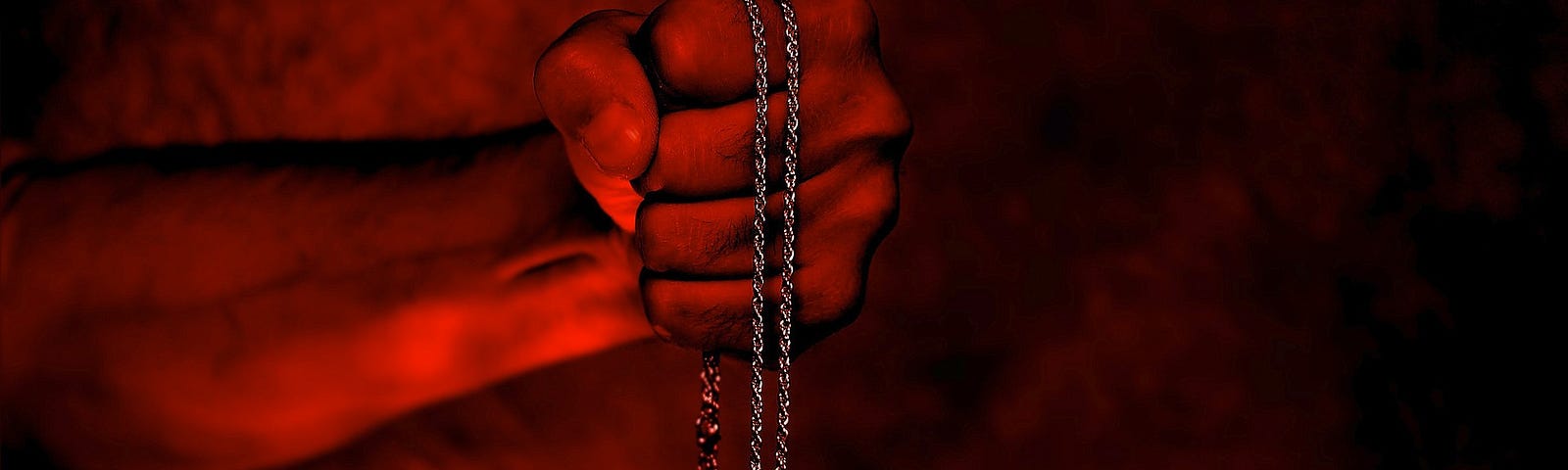 A red-tinted photo manipulation of a hand holding a silver chain with a heart-shaped pendant covered with red gems.