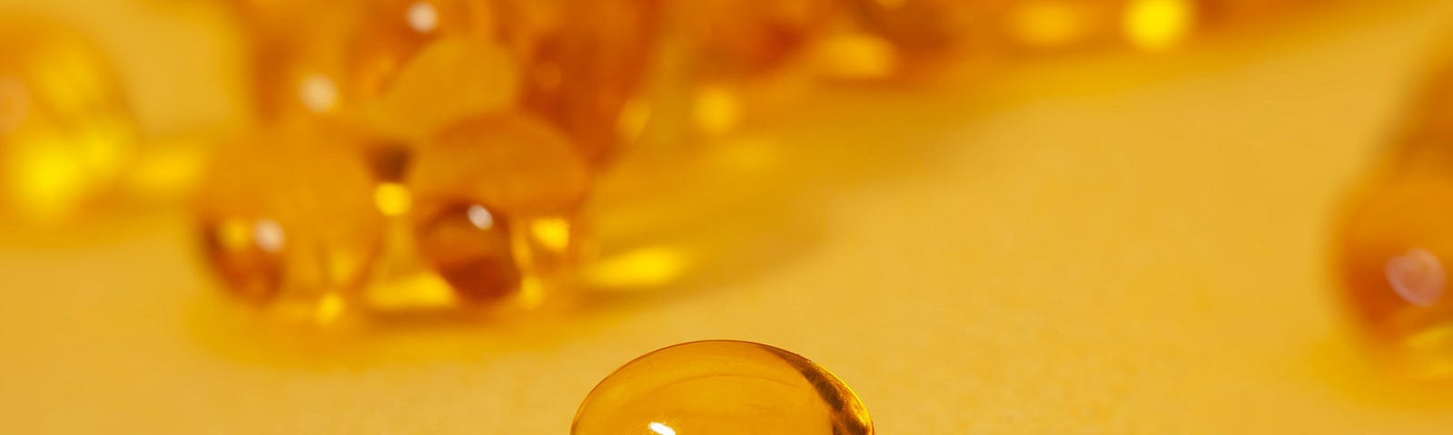 A yellow vitamin gel pill with several more of them blurred against a yellow background.