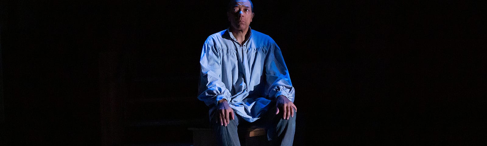 A photograph of a man with brown skin and short dark hair. He wears a light blue shirt with billowing sleeves over gray pants, and sits in a chair alone in a jail cell.