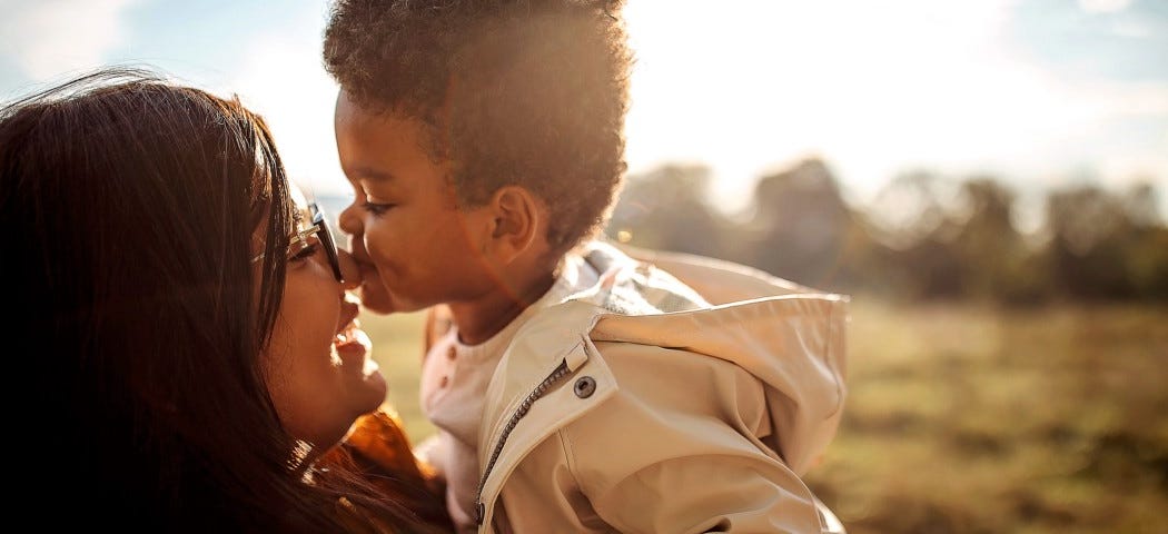 Un niño pequeño besando a su madre en la nariz mientras ésta lo abraza. Están de pie en la naturaleza contra la luz del sol.
