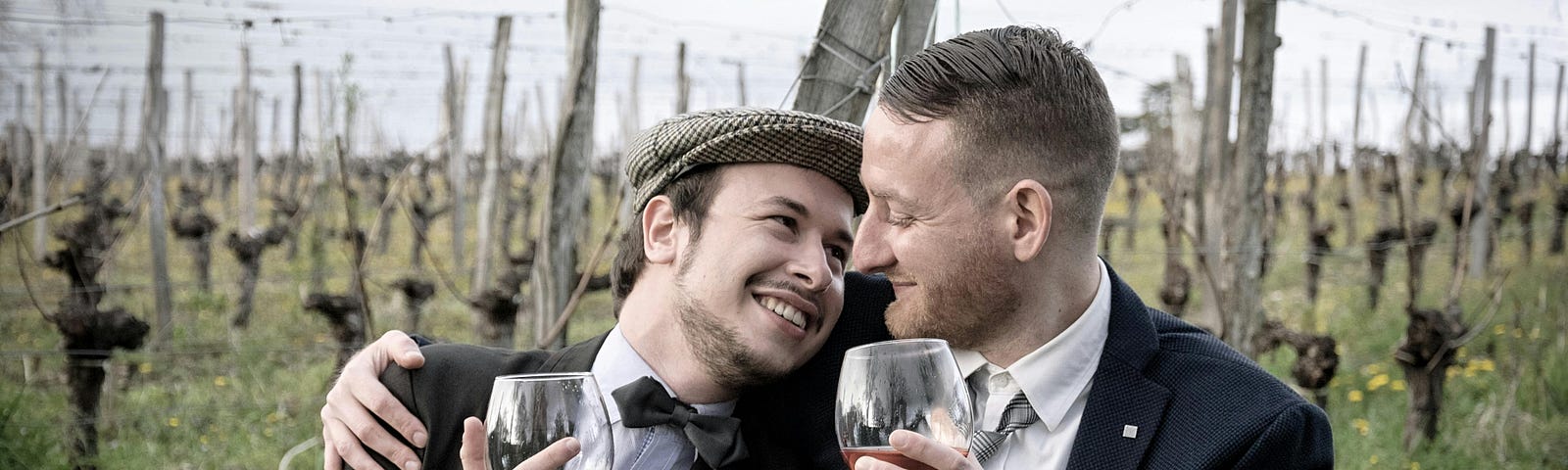 Two men holding wine glasses in a field, cuddled together and staring lovingly into each other’s eyes