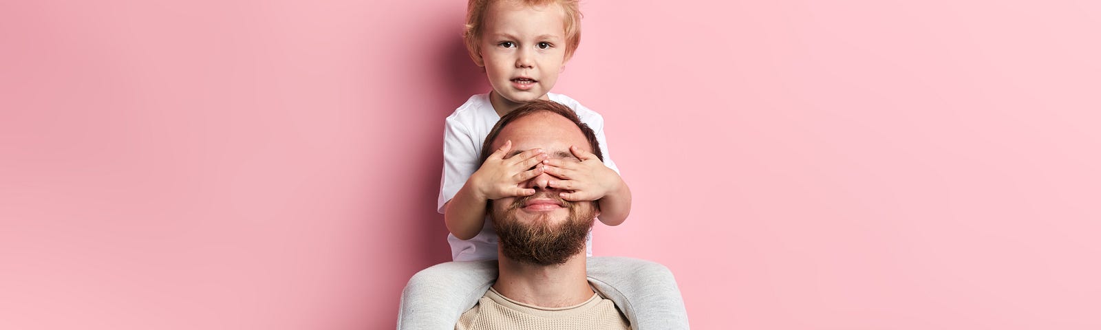 little boy closing his dad’s eyes with palms, close up portrait, isolated pink background, studio shot