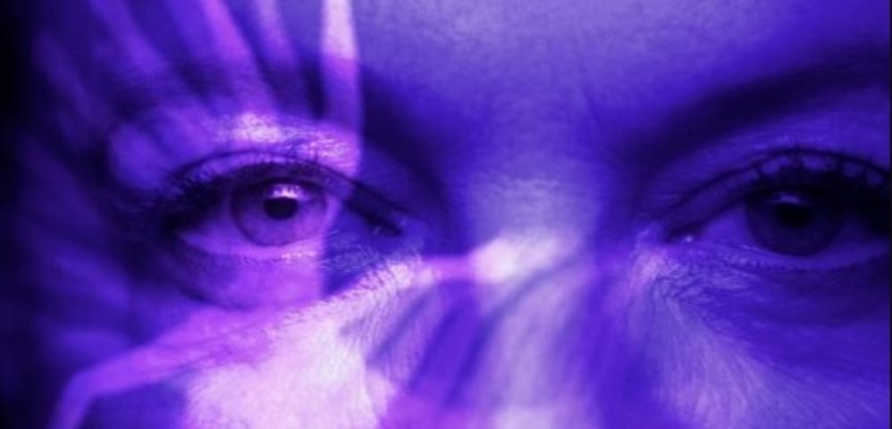 A closeup of a white woman’s eyes. A vague film image projects across her face, coloring it blue.