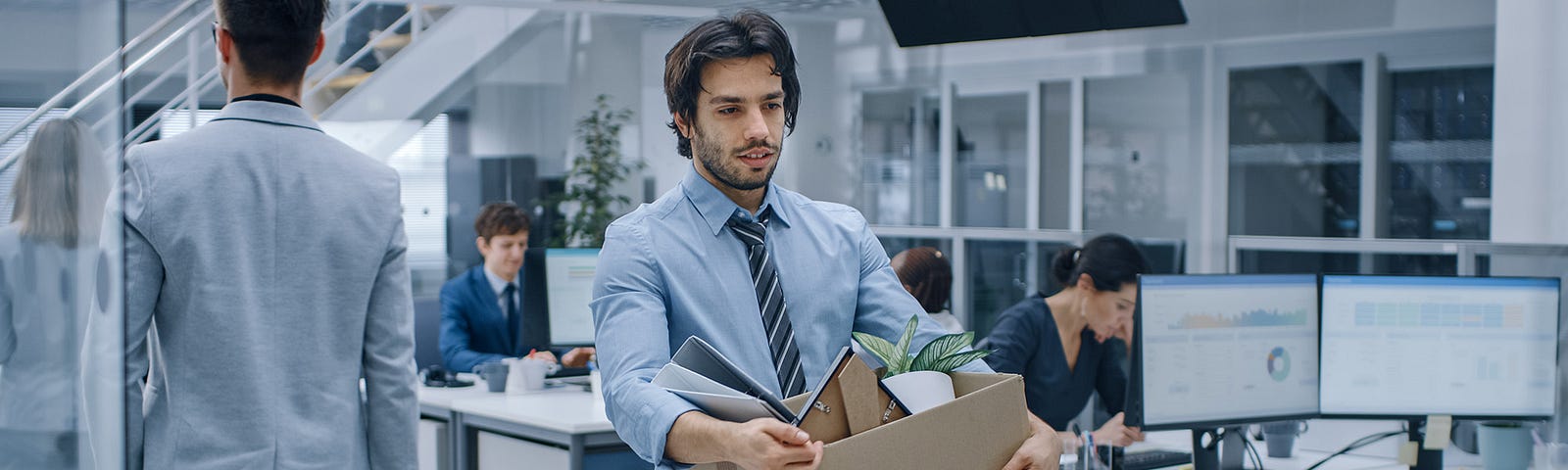 Man wearing tie in office walking out with box in arms after just being laid off